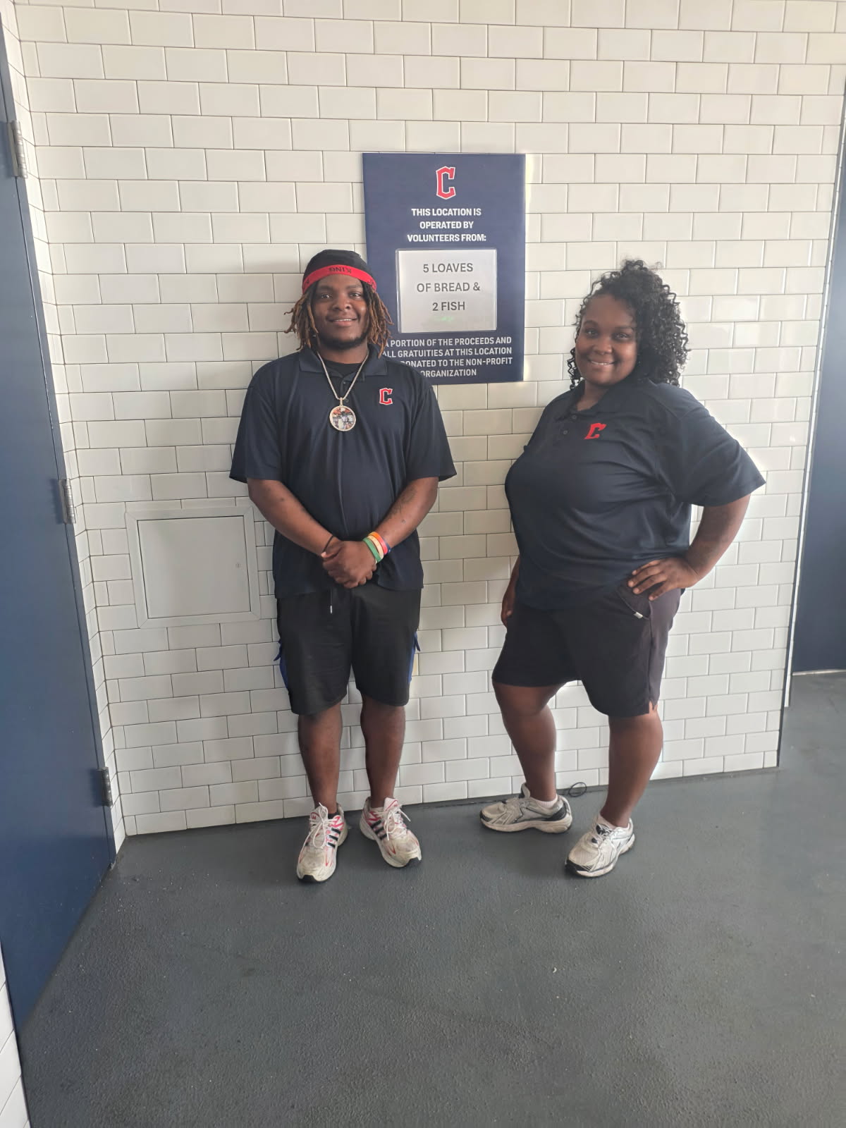 Two smiling 5 Loaves 2 Fish volunteers in navy polo shirts in front of the stadium sign