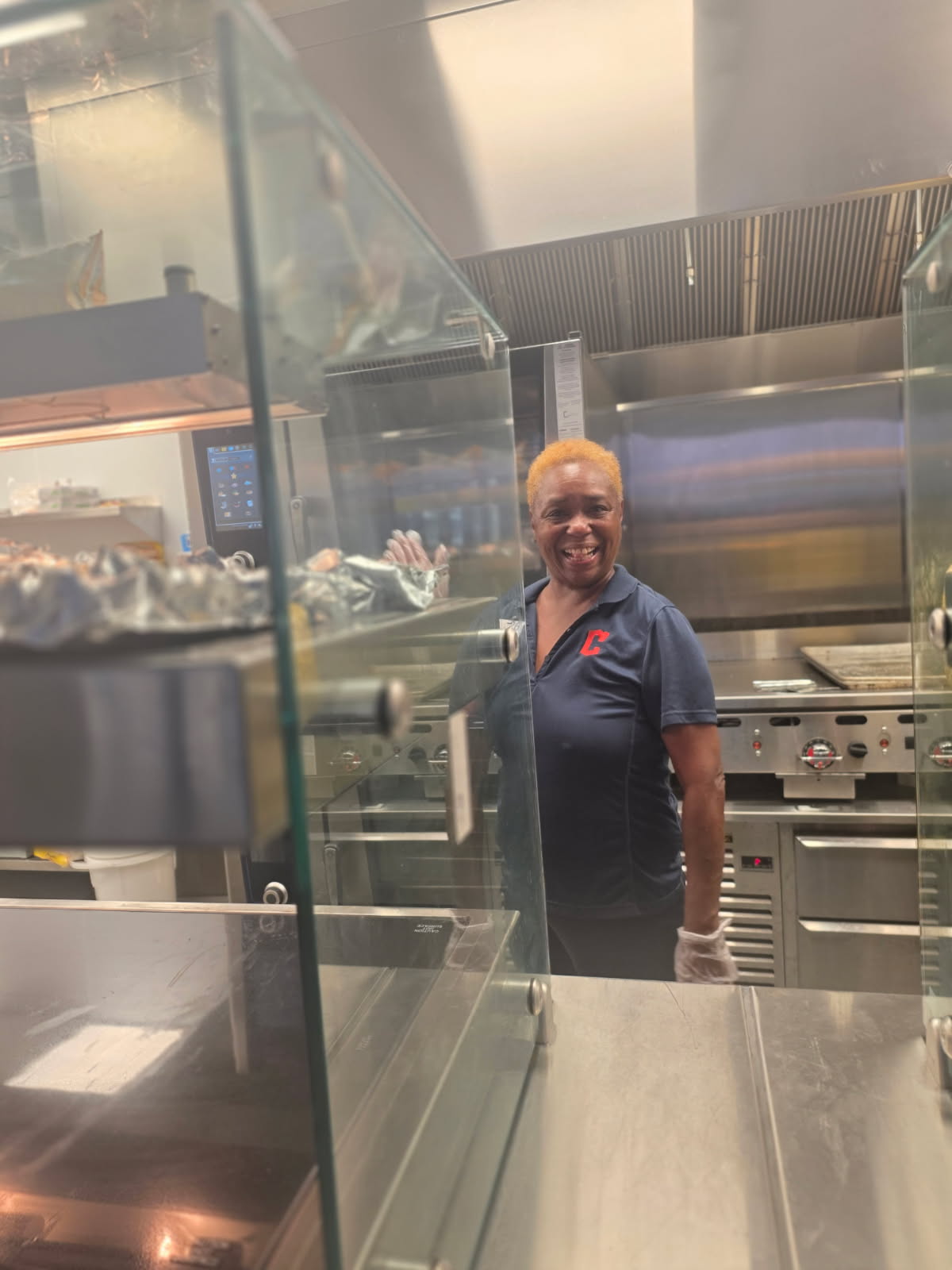 A smiling 5 Loaves 2 Fish volunteer in a navy polo with a red C logo, working in the stadium concession kitchen